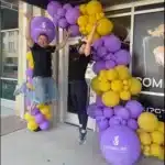two people dressed in black, jumping in front of store with an Upgrade Labs sign, with a purple and yellow balloon arch